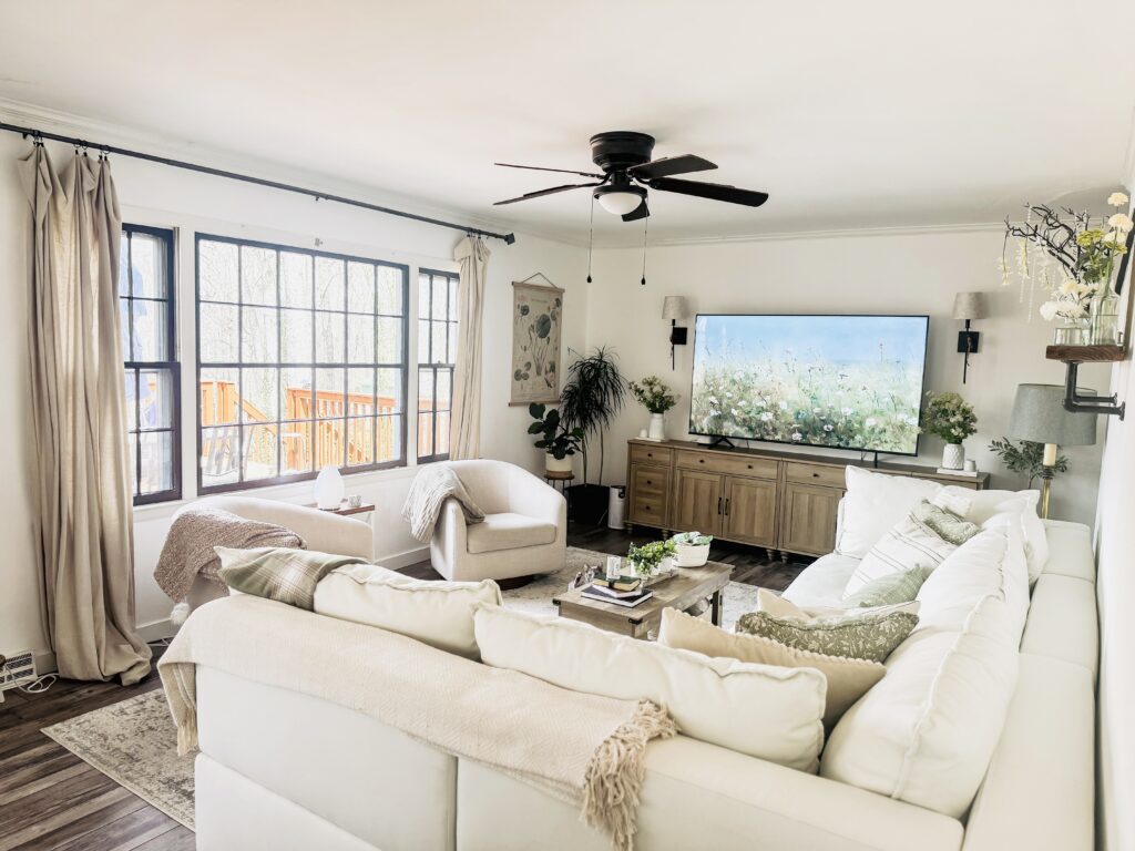 Bright and inviting living room featuring large windows, comfortable white sofas, a wooden TV console, and lush greenery for a relaxing home atmosphere.