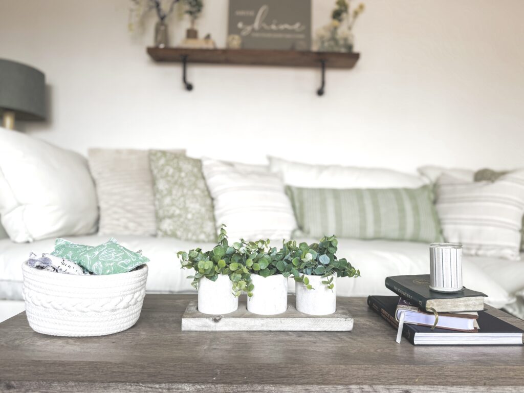 A cozy living room featuring a wooden coffee table with potted green plants, a basket of textiles, and a background of white and green cushions on a sofa, creating a relaxing and inviting atmosphere.