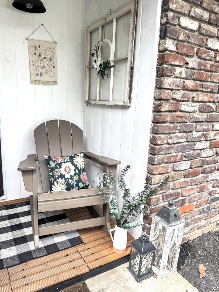 A charming outdoor corner featuring a beige Adirondack chair with floral cushion, potted greenery, vintage window frame, and lanterns on a wooden porch with brick wall background.