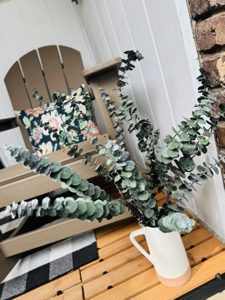 A beautiful arrangement of eucalyptus branches in a white ceramic vase, placed on a wooden porch floor near a cozy outdoor seating area with floral cushions.
