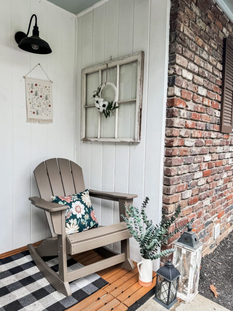 A cozy porch corner featuring a beige Adirondack chair with floral cushion, a white window frame with a floral wreath, and decorative lanterns, creating a welcoming outdoor space.