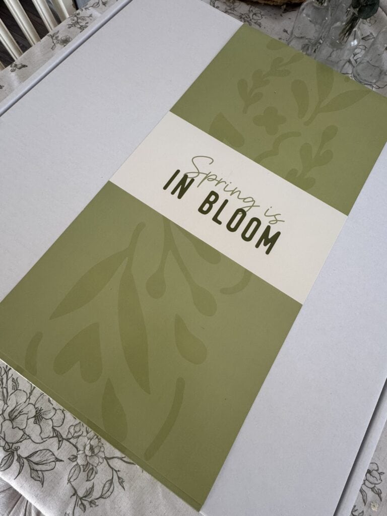 Decorative spring-themed sign with the message "Spring is in Bloom" in elegant script and bold lettering, perfect for spring decor and floral arrangements.