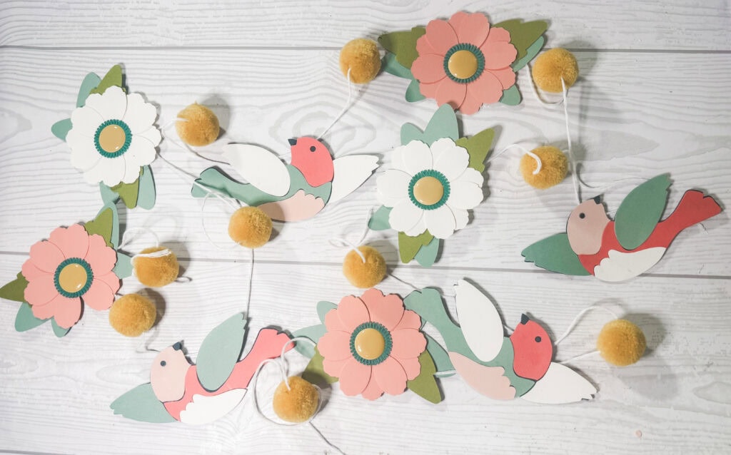 Colorful paper flower and bird garland with yellow pom-pom accents, perfect for spring or Easter decor, crafted from paper and string for a charming, festive look.