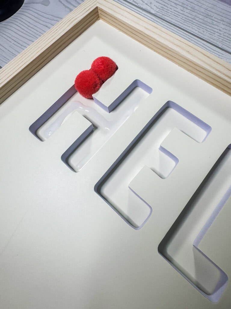 Decorative love sign with white cut-out letters and red heart-shaped pom-poms, placed on a white surface with a wooden frame, perfect for romantic or wedding-themed decor.