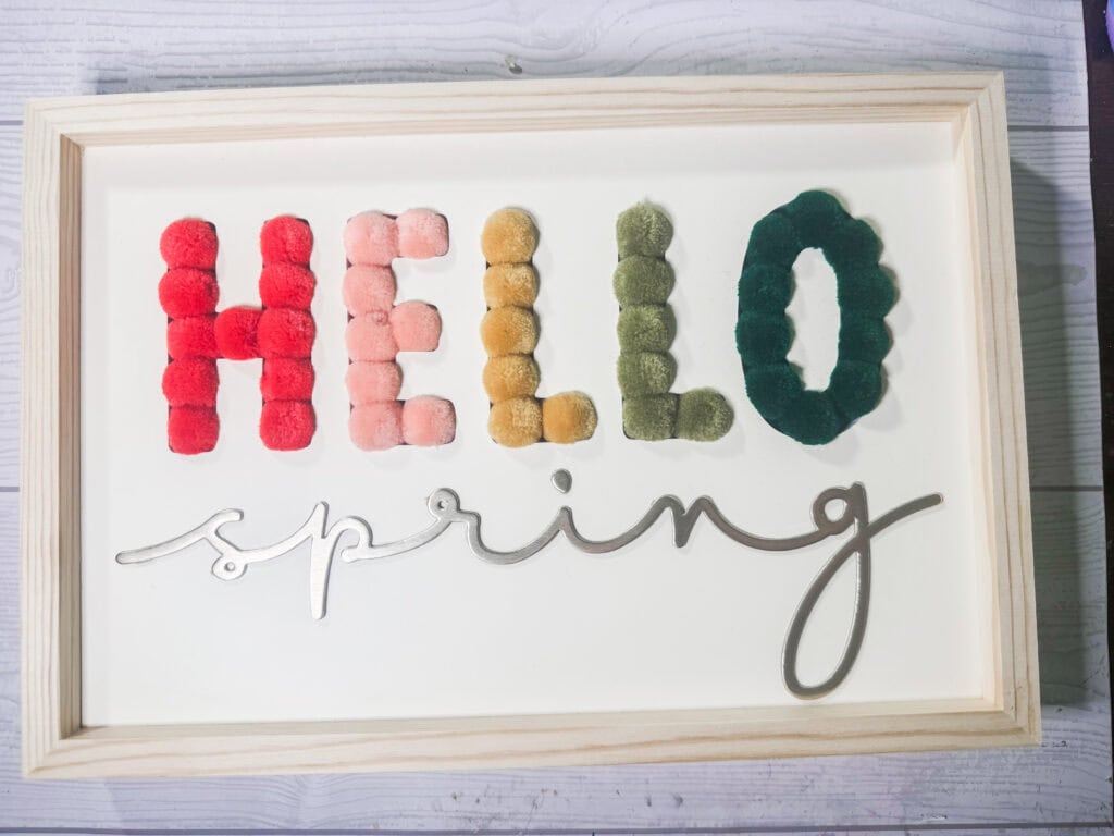 Colorful spring-themed sign with pom-poms spelling "HELLO" in vibrant red, pink, yellow, green, and dark green, with a white background and elegant "spring" script, perfect for seasonal decor.