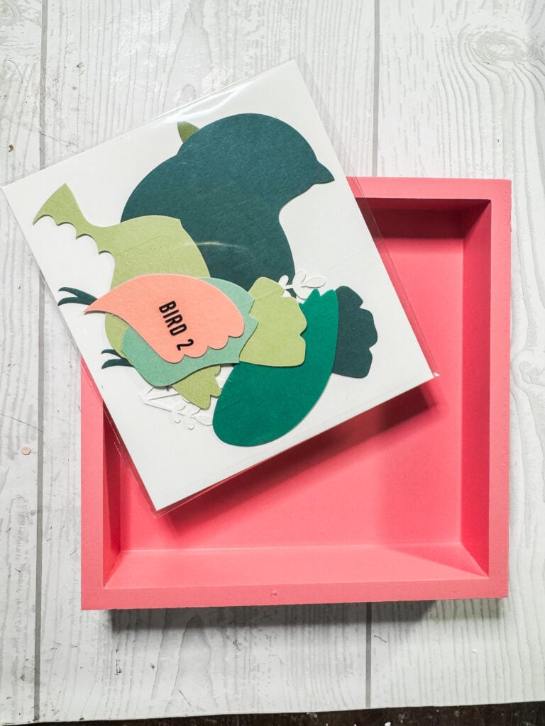 Colorful layered paper bird craft for kids, featuring a bird cutout with a "BIRD 2" label, displayed in a pink box on a white wooden surface.
