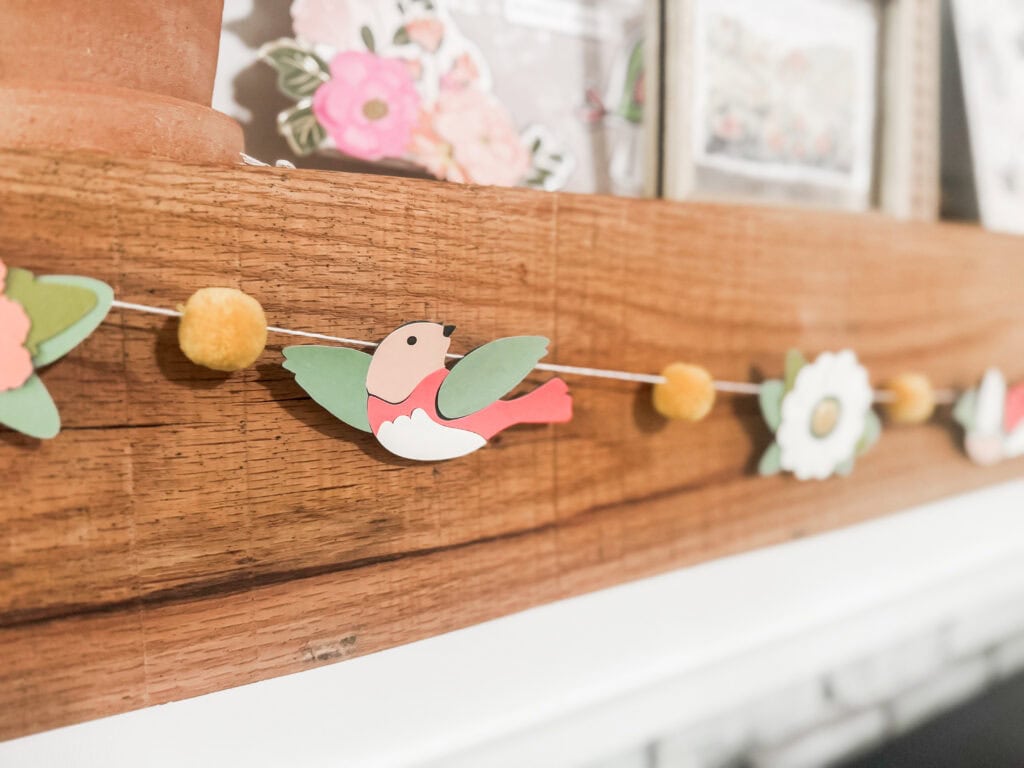Decorative bird and flower garland made of paper and felt, hanging on a wooden surface, ideal for spring or garden-themed decor, perfect for adding a charming touch to any space.
