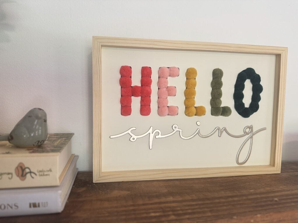 Bright spring-themed "HELLO" sign with colorful yarn letters and elegant "spring" script, perfect for seasonal home decor or greeting displays.