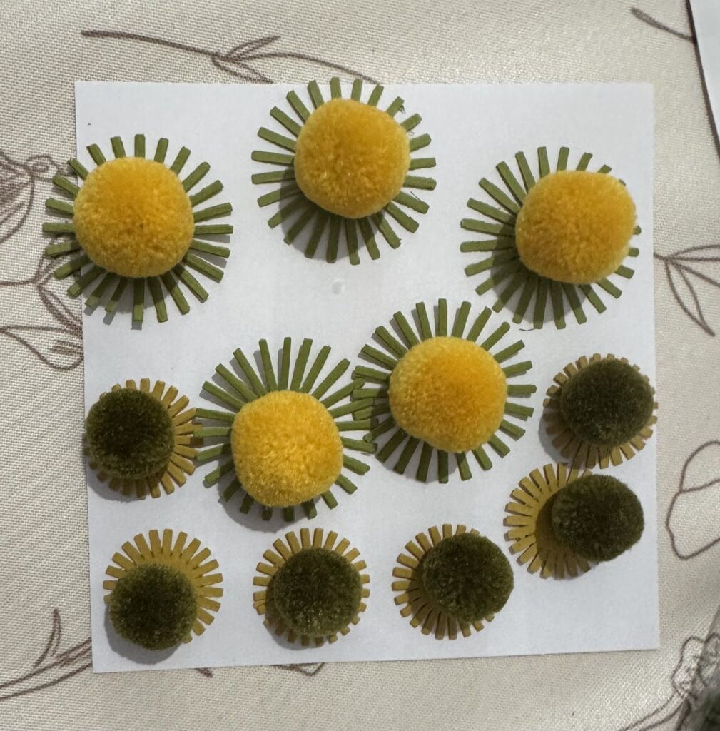 Bright yellow and dark green pom-poms with green and yellow paper sunburst accents, arranged on a white background for decorative craft or DIY projects.
