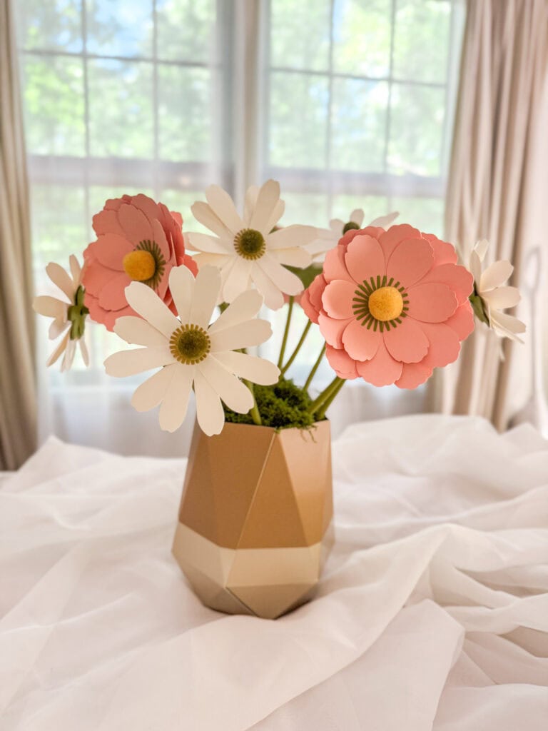 A charming paper flower bouquet featuring pink and white paper daisies arranged in a modern geometric vase, set on a soft white fabric with a bright window background.