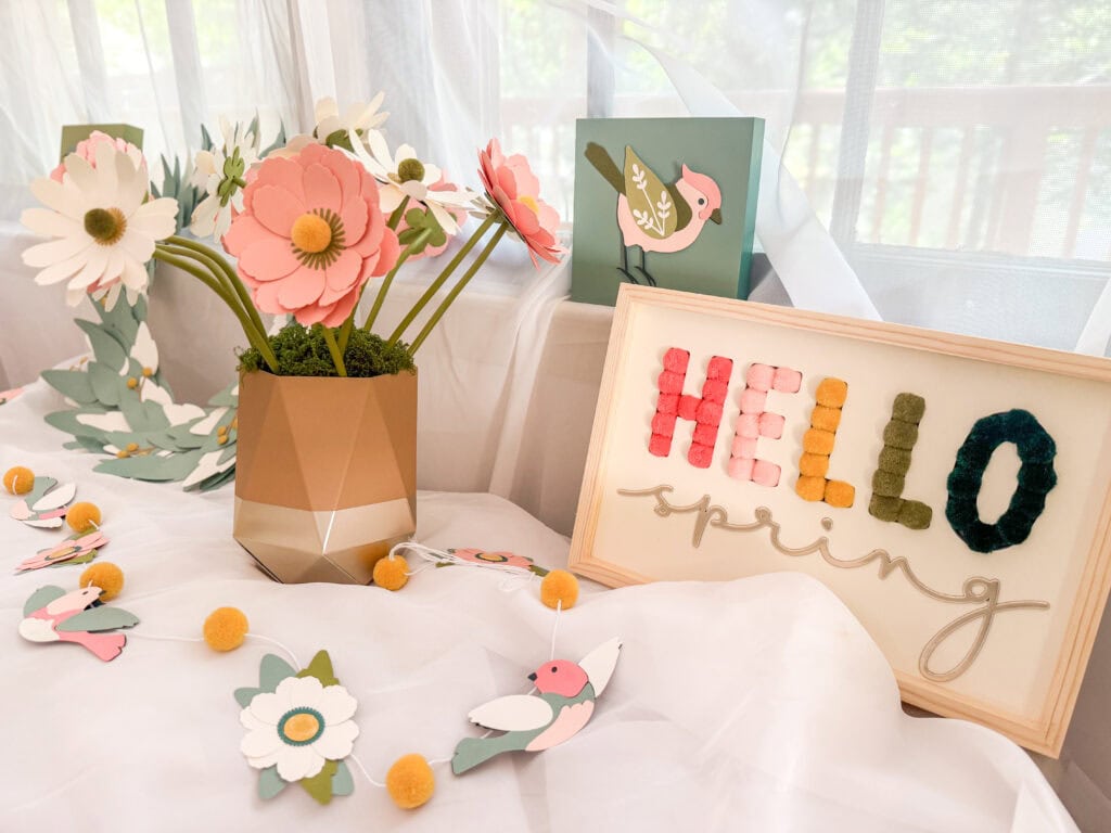 Bright spring-themed decor featuring paper flowers, a framed "Hello Spring" sign, and bird illustrations, perfect for celebrating the season with cheerful and colorful accents.