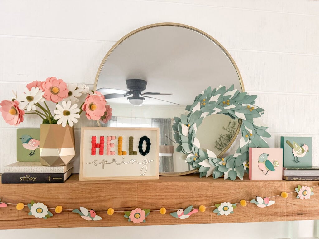 Bright spring-themed decor featuring a floral arrangement, bird illustrations, and a cheerful "HELLO spring" sign on a wooden mantel with a round mirror.