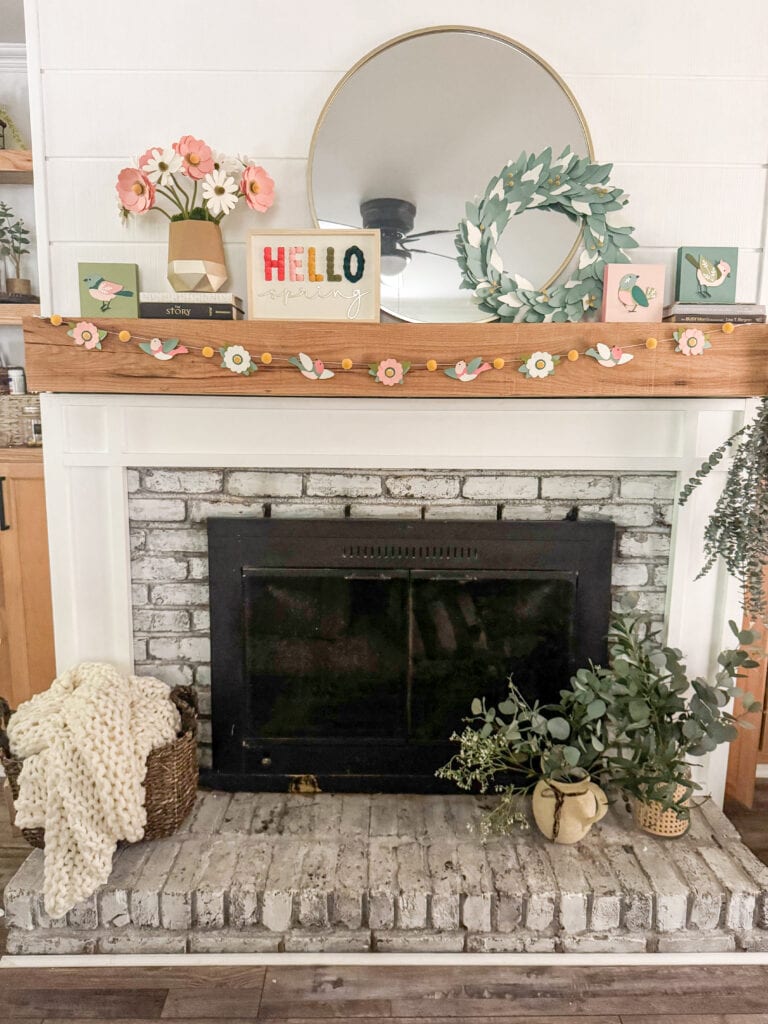 A cozy fireplace mantel decorated with pink and white flowers, greenery, and charming artwork, creating a warm and inviting home decor setting.