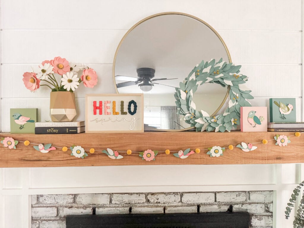A cozy spring-themed mantel decorated with pink and white flowers, a round mirror, bird-themed accents, and a "HELLO spring" sign, creating a warm and inviting home atmosphere.