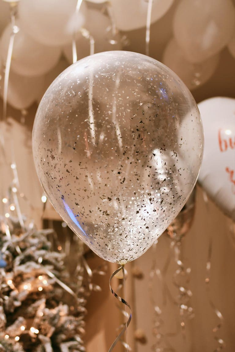 A large transparent balloon filled with silver confetti, perfect for festive occasions and celebrations, surrounded by other balloons and holiday decorations.