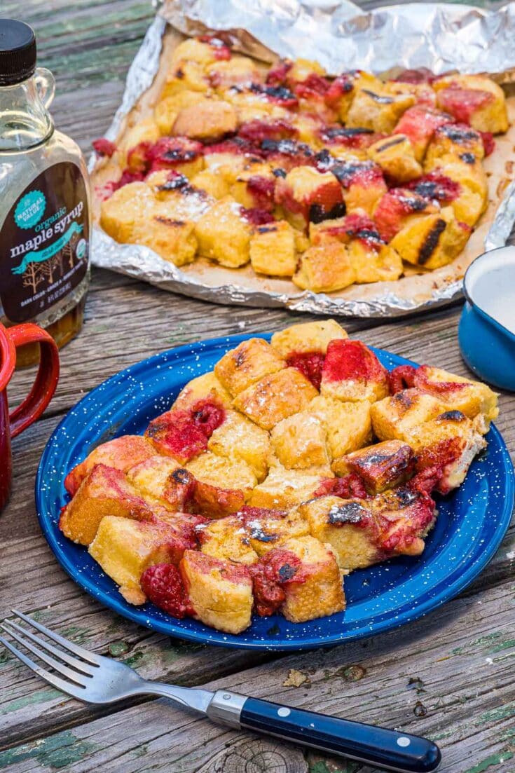Delicious berry cobbler with fresh raspberries and blackberries, topped with golden, flaky biscuit crust, served on a blue plate with a fork, outdoors on a rustic wooden table.