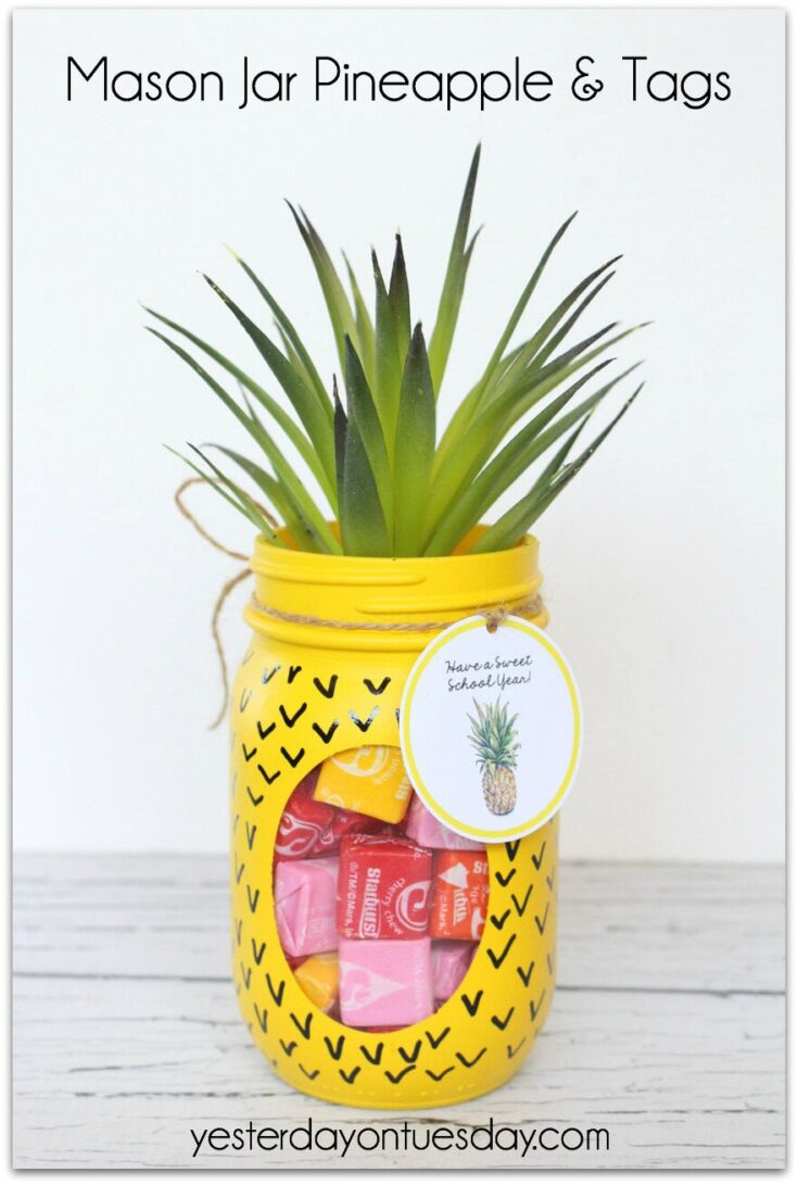 Bright yellow mason jar decorated with black chevron patterns, filled with pink and red candies, topped with a real pineapple, and labeled with a pineapple tag for a gift or decor.