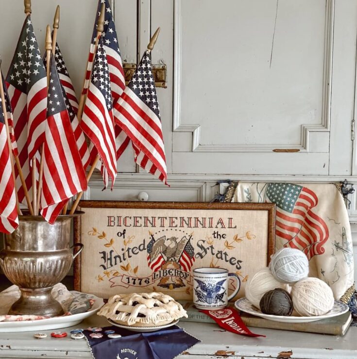 An arrangement of small American flags in a vintage metal holder, complemented by patriotic-themed decor including yarn balls, a "Bicentennial of the United States" framed print, and a patriotic mug,.