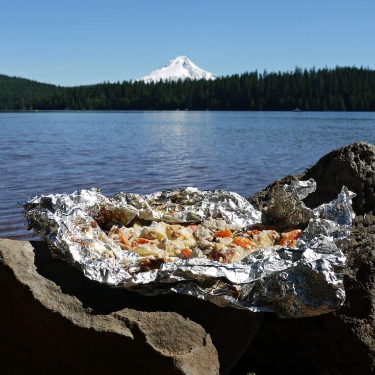 A foil-wrapped pizza on a rock by a lake, with a snow-capped mountain and forest in the background, under a clear blue sky.