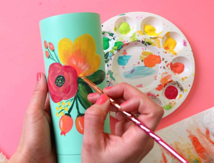 A person is painting a vibrant floral design on a ceramic mug using a fine brush and acrylic paints, with a paint palette nearby, on a pink surface.
