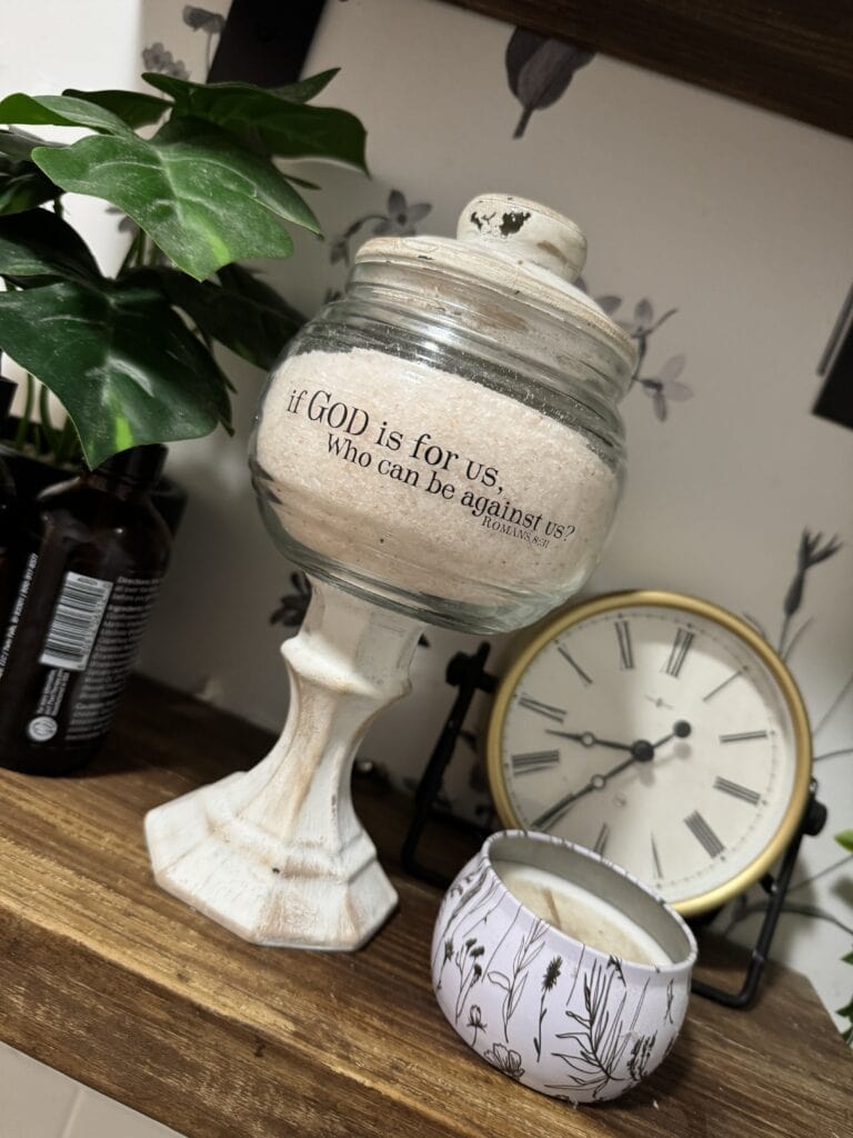 Decorative glass jar with a biblical quote, placed on a wooden surface alongside a potted plant, clock, and candle, creating a cozy and inspirational home decor scene.