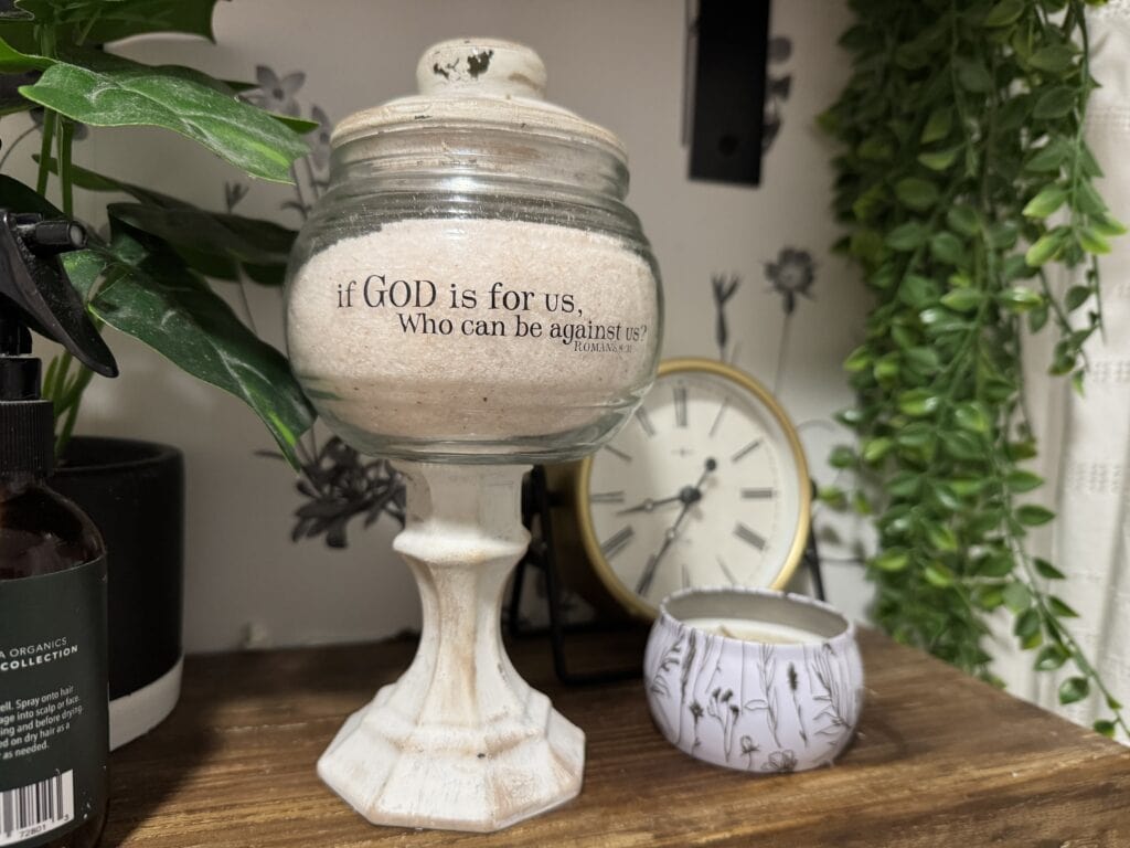 Decorative ceramic jar with the quote 'if GOD is for us, Who can be against us?' on a wooden surface, surrounded by plants and a clock in the background.