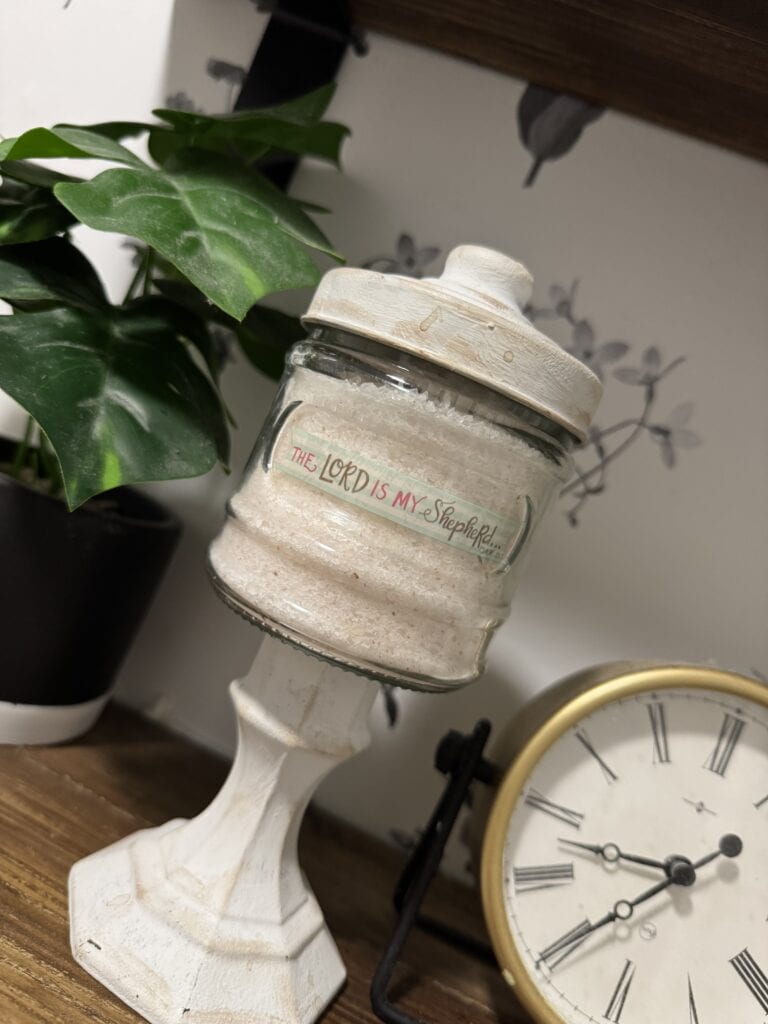 A decorative candle holder with a religious message, placed on a wooden surface next to a clock and a potted plant, creating a cozy and spiritual ambiance.
