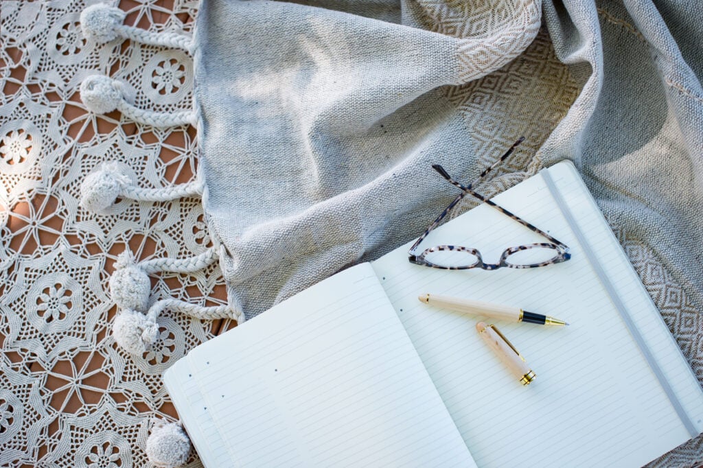 A flat lay of an open notebook, reading glasses, and a pen on a textured fabric surface, promoting mindful and slow living practices.