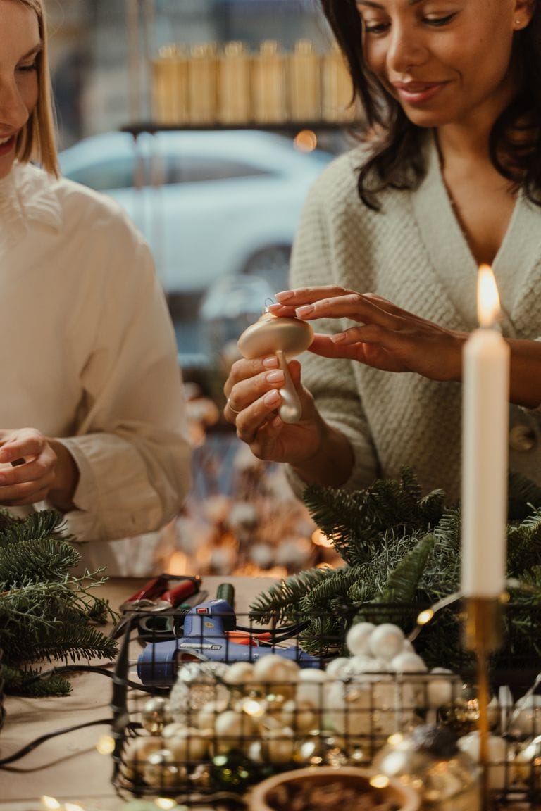 Two women lighting candles during a festive holiday celebration, surrounded by Christmas decorations, candles, and greenery, creating a warm and joyful atmosphere.