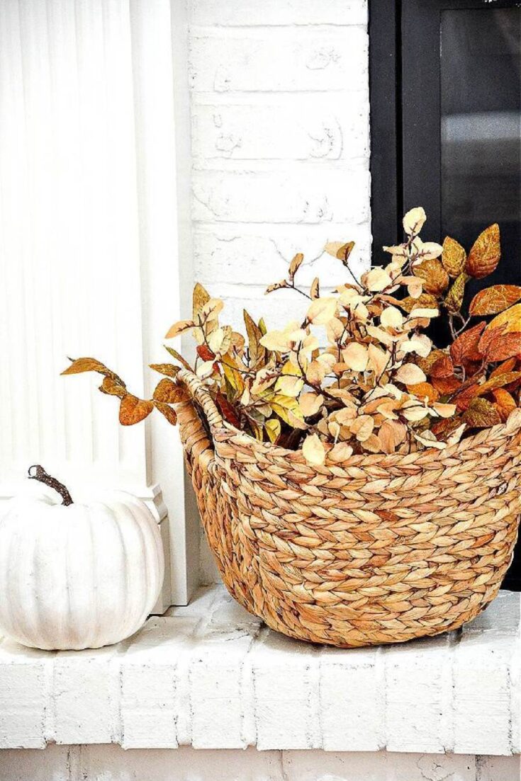 A woven basket filled with dried autumn leaves and branches, placed on a white brick ledge next to a white pumpkin, creating a cozy fall decor scene.