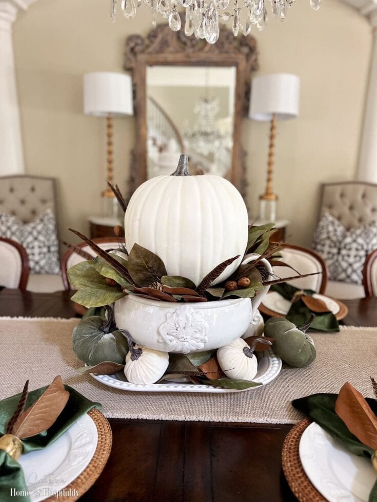 Stylish fall table centerpiece featuring a large white pumpkin surrounded by greenery and small pumpkins, set in a sophisticated dining room with a chandelier and vintage mirror.