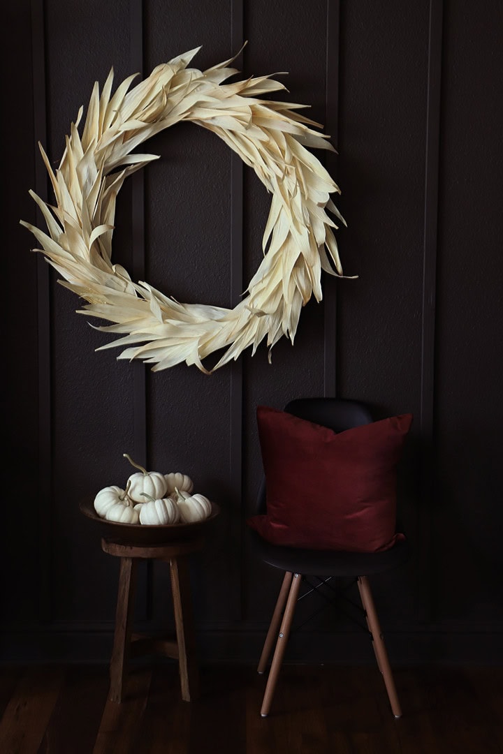 Elegant fall wreath made of dried leaves hanging on a dark wall, with a small table holding white pumpkins and a cozy red pillow, perfect for autumn home decor.