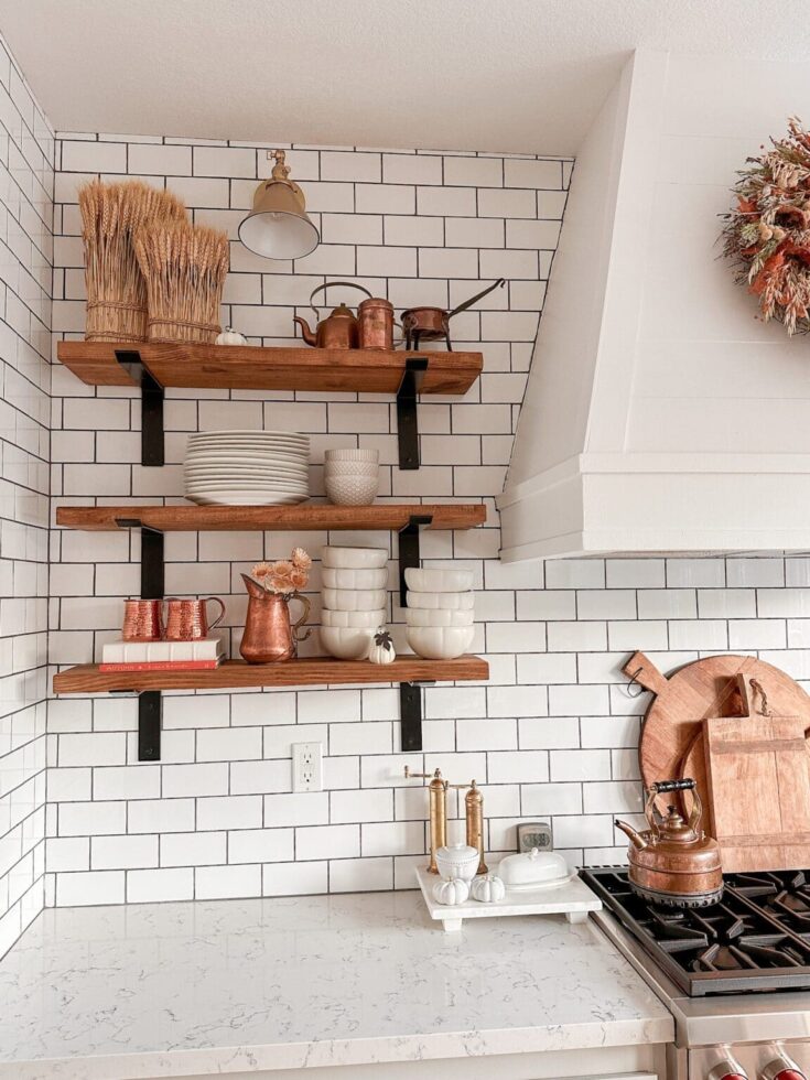 Rustic fall kitchen decor with wooden shelves, white subway tile backsplash, copper accents, and seasonal wreath for cozy autumn ambiance.