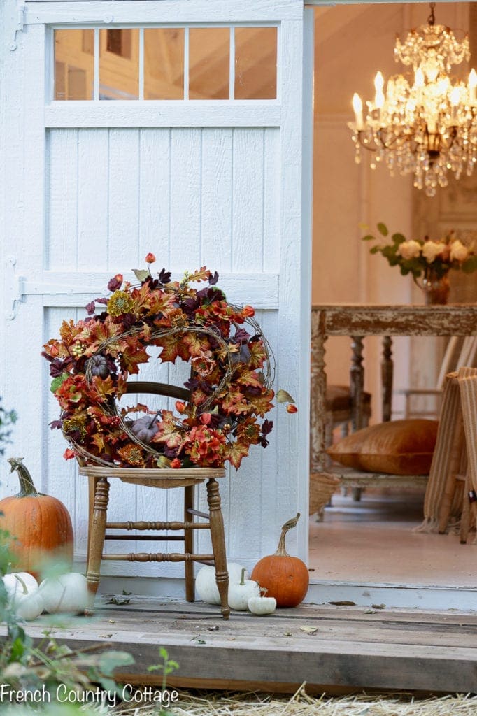 A fall-themed wreath with colorful leaves and berries on a wooden chair outside a cozy cottage, with pumpkins and gourds nearby, and a glimpse of an elegant chandelier inside.