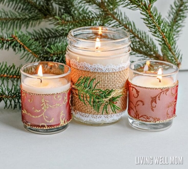 Three lit candles in decorative glass holders with pink and gold patterns, surrounded by pine branches, creating a cozy holiday ambiance.