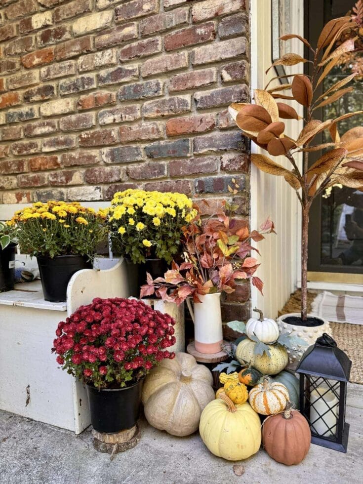 Autumn porch decor featuring pumpkins, mums, and fall foliage against a brick wall, perfect for a cozy fall home decorating idea.