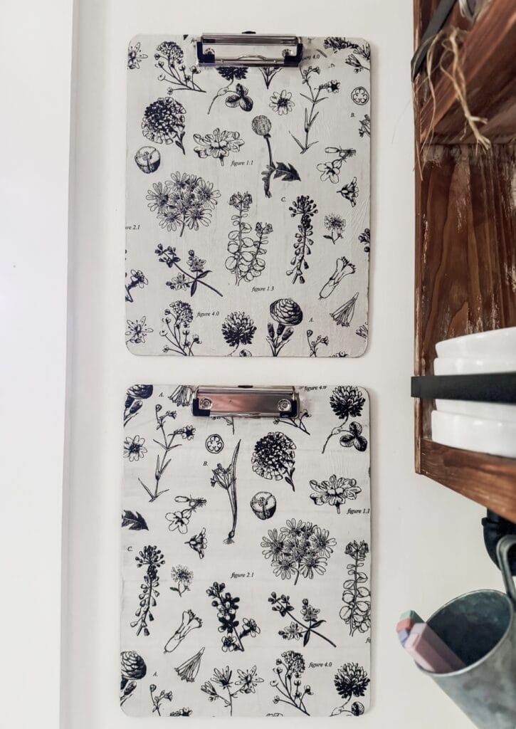 Two clipboards with black and white botanical illustrations of various flowers and plants, mounted on a white wall, with a wooden shelf and kitchen utensils nearby.