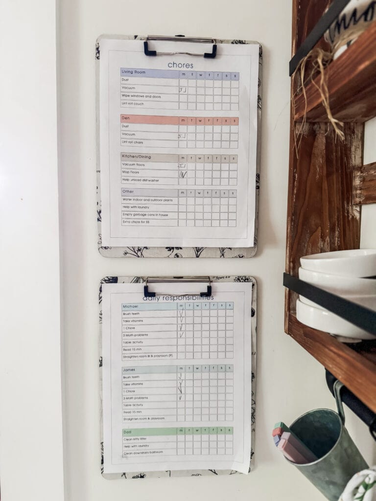 Two whiteboards with chore and daily responsibilities checklists, mounted on a white wall in a home setting, with shelves holding dishes and kitchen utensils nearby.