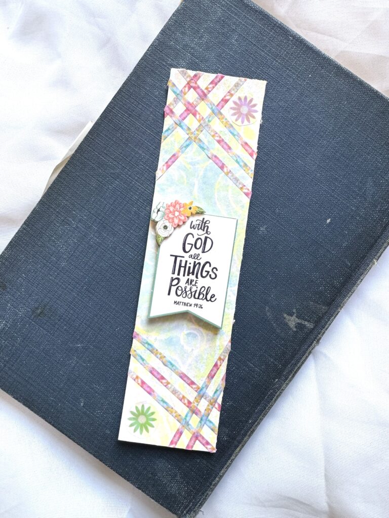 Colorful bookmark with a faith-based quote from Matthew 19:26, placed on a black book with a white fabric background, promoting hope and belief in the impossible.