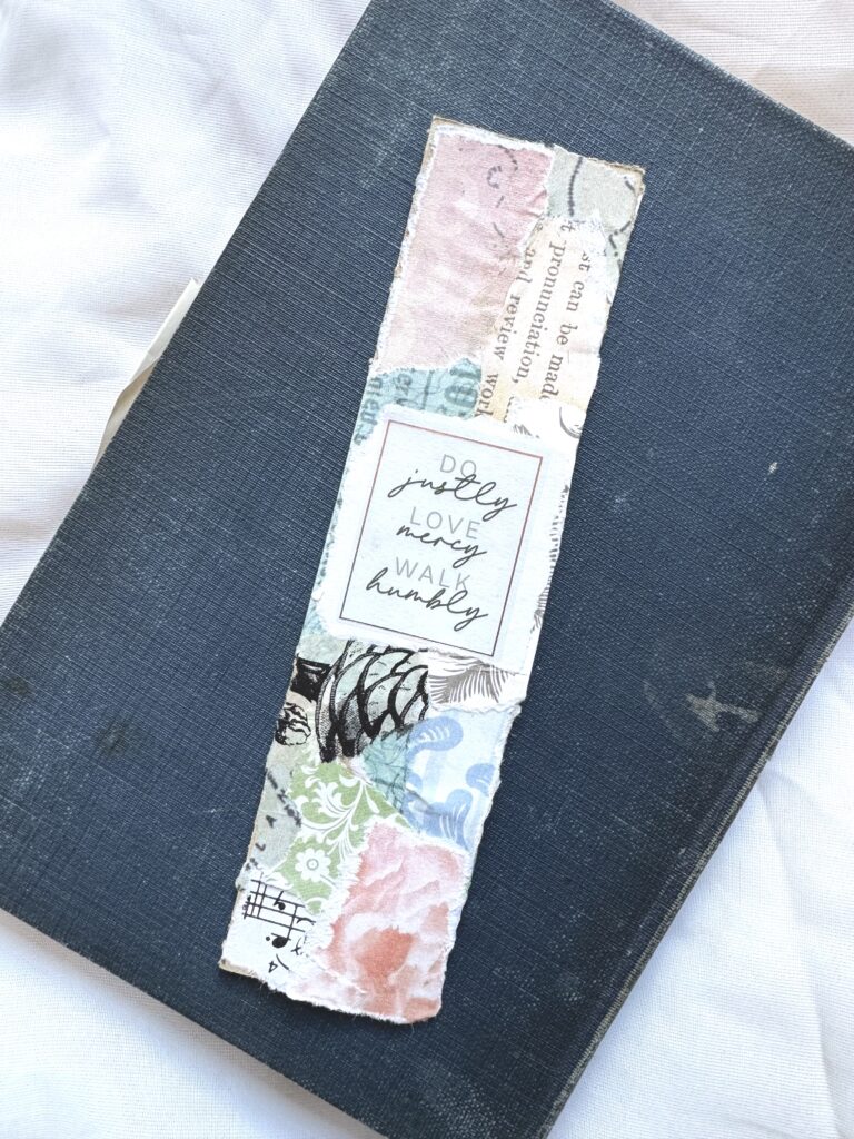 A handmade decorative bookmark with a handwritten quote about love, joy, and walking humbly, placed on a dark textured book cover for aesthetic appeal.