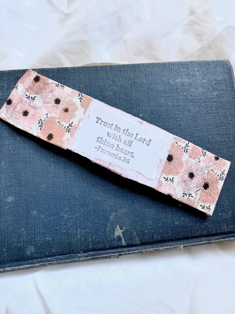 A Bible verse bookmark with pink floral pattern and Proverbs 3:5 quote, placed on a dark blue book, for spiritual and inspirational decor.