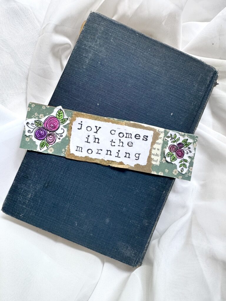 A blue hardcover notebook with a decorative band featuring purple roses and the quote 'joy comes in the morning' for uplifting and motivational content.