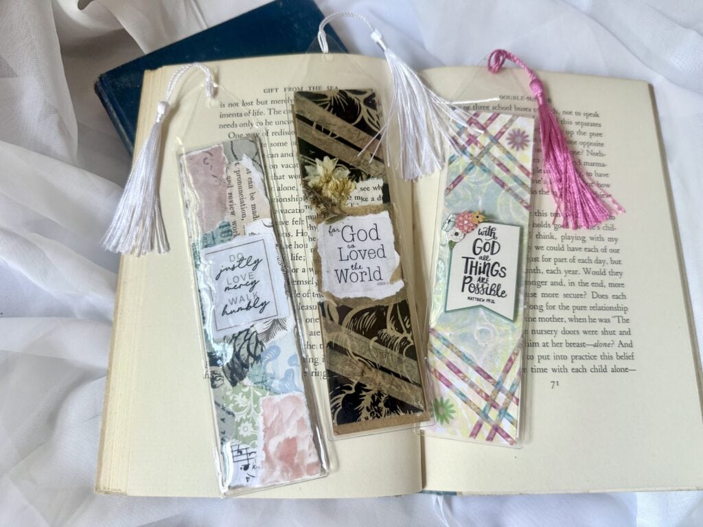 Set of three colorful bookmarks with tassels and inspirational messages, placed on an open book, perfect for Bible journaling and reading accessories.