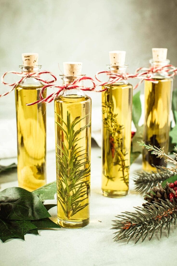 Four glass bottles filled with herbal infused olive oil, decorated with red twine, surrounded by pine branches and holly leaves, on a light background.