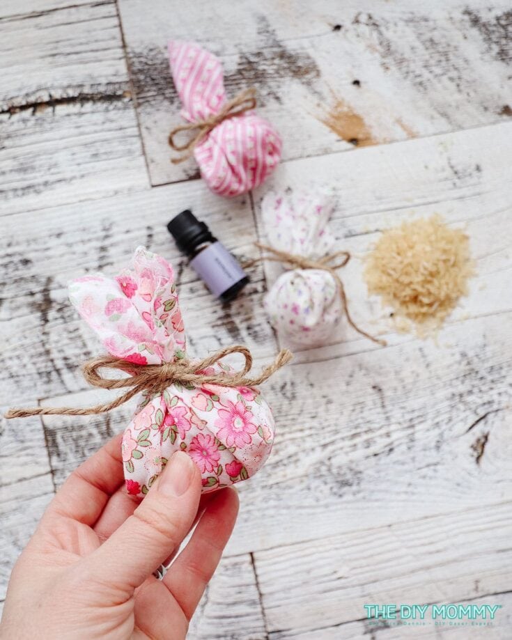 Hand holding a small fabric lavender sachet with other sachets, essential oil bottle, and natural filling on rustic wooden background for homemade gift ideas.