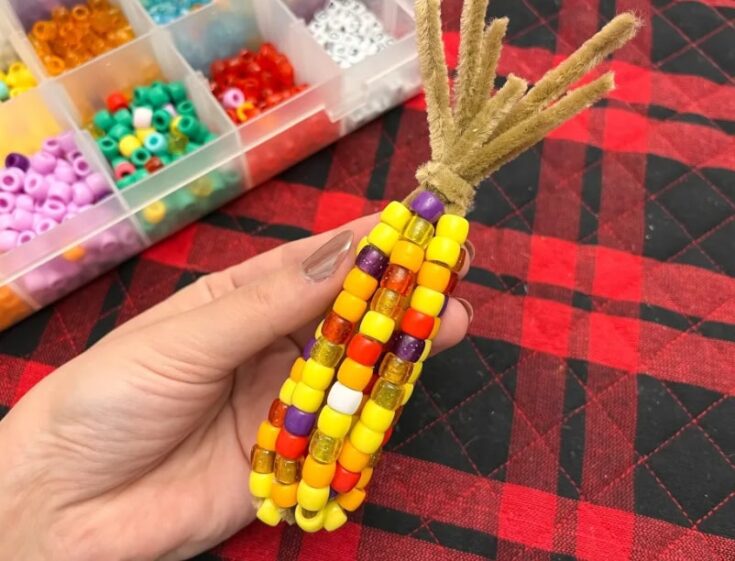 Hand holding a beaded corn craft made with colorful beads and yarn, on a red and black checkered fabric background, with a container of beads in the background.
