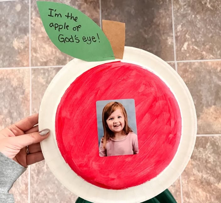 Colorful craft project featuring a paper apple with a child's photo and a green leaf with the phrase "I'm the apple of God's eye!" for children's religious activities.