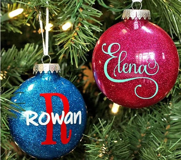 Two glittery Christmas ornaments hanging on a tree, one blue with a red letter D and the name Rowan, and one pink with the name Elena, celebrating holiday cheer.