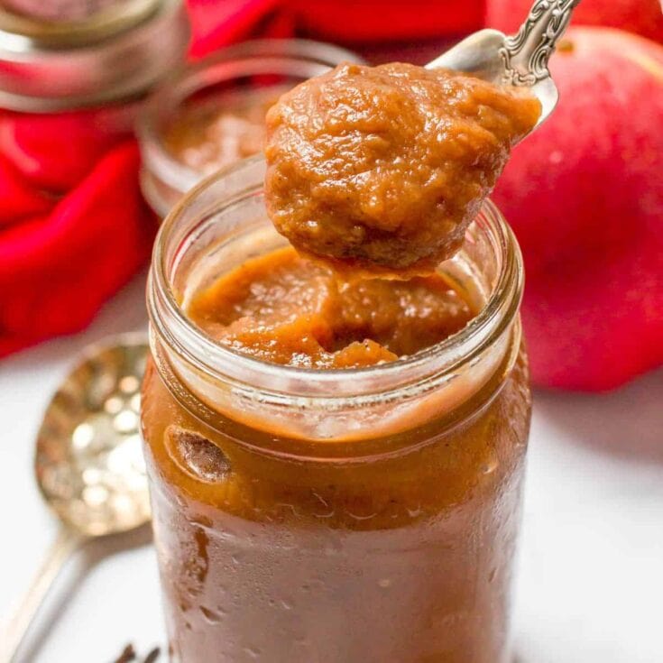 Close-up of a spoonful of homemade apple butter above a glass jar filled with apple butter, with fresh apples in the background, on a white surface.