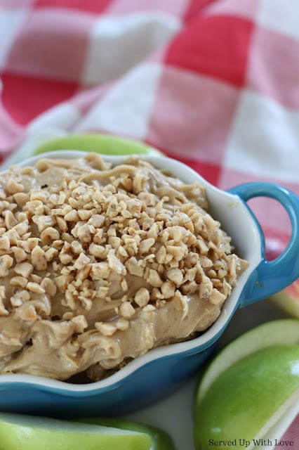 Close-up of apple crisp topped with chopped nuts in a blue baking dish, with fresh green apples and a red and white checkered cloth in the background.